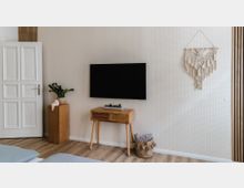 Heller Wohnraum mit einer weißen Tür, einer modernen gemusterten Tapete und einem Wandfernseher. Darunter steht ein kleiner Holztisch mit Pflanzen darauf, daneben ein Korb aus Stoff und ein Makramee-Wandbehang.