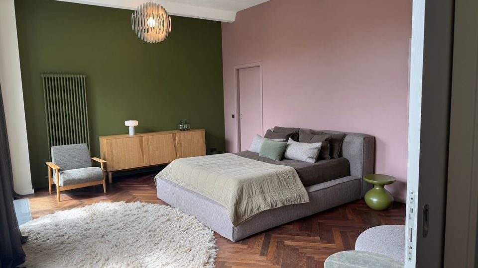 Modern bedroom with a low upholstered bed, green accent wall and pink side wall, and herringbone wood flooring. The room includes pendant lights, a wooden sideboard, an armchair, a round shag rug, and a small green side table.