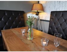 Ein Holztisch in einem Restaurant mit zwei gepolsterten, dunkelbraunen Lederbänken im Hintergrund. Auf dem Tisch stehen eine Vase mit weißen Blumen, zwei Teelichter in Glasbehältern und ein leeres Weinglas; daneben eine Stehlampe mit braunem Lampenschirm.