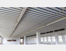 Großer Innenraum mit Industriecharakter, gekennzeichnet durch eine abgehängte Decke mit blauen Streifen und langen Leuchtstofflampen. Die Wände sind weiß, mit mehreren rechteckigen Fenstern und vereinzelten weißen Säulen, die den Raum gliedern.