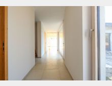 Heller Flur in einem modernen Gebäude mit beigen Wänden und Bodenfliesen, links eine offene Holztür und rechts ein großes Fenster mit Holzrahmen. Am Ende des Flurs befindet sich ein heller Raum mit Glasfenstern.