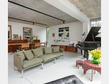Modern open-plan living and dining room with exposed concrete ceiling, white walls, and large windows, featuring a green modular sofa on a light rug and a black grand piano. A long wooden dining table with chairs, low wood cabinets, and framed artwork line the walls, with a small red side table holding a vase of yellow flowers.