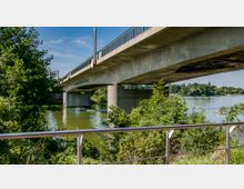 Eine moderne Betonbrücke überspannt einen Fluss, umgeben von grünen Bäumen und Sträuchern. Im Vordergrund ist ein Geländer zu sehen, während unter der Brücke ein kleines Boot auf dem Wasser fährt.
