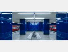 Eine U-Bahn-Station mit glänzenden blauen Wänden und Säulen. In der Mitte befindet sich eine Sitzbank, und an den Seiten sind rote Paneele mit weißen und blauen Linien sowie der Aufschrift „Osloer Straße“ sichtbar.