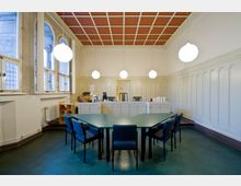 Ein historischer Besprechungsraum mit hohen Decken, verzierten Wandpaneelen und großen Bogenfenstern. Im Vordergrund steht ein grüner Konferenztisch mit gepolsterten Stühlen, während im Hintergrund ein Empfangstresen mit Computern und hängenden Kugelleuchten sichtbar ist.