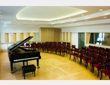 Ein moderner Konzertsaal mit Holzboden, einem schwarzen Flügel im Vordergrund und einer halbkreisförmigen Anordnung roter Polsterstühle. Die Wände sind hell mit integrierten Holzpaneelen und großen Fenstern.