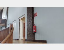 Ein Treppenaufgang in einem Innenraum mit Holzgeländer, einer hellgrauen Wand und einer gemauerten Säule. An der Wand befindet sich ein Feuerlöscher unter einem entsprechenden Hinweisschild; daneben sind geöffnete weiße Türen zu sehen, die in einen weiteren Raum führen.