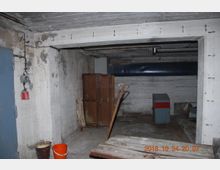 Kellerraum mit unverputzten Betonwänden und Decke, darin alte Werkzeuge, Holzkisten und ein rostiger Palettenhubwagen. Im Vordergrund ein orangefarbener Eimer und ein Metalleimer, im Hintergrund ein großer Metallschrank und Belüftungsrohre.