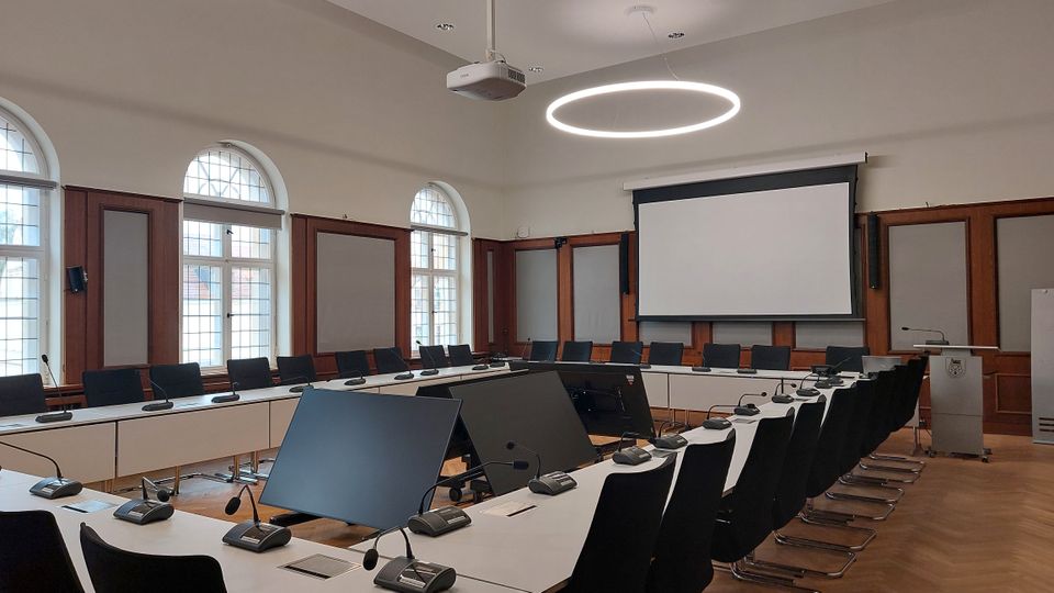 Moderner Konferenzraum mit hufeisenförmiger Anordnung von weißen Tischen und schwarzen Stühlen. Der Raum verfügt über große Fenster mit Rundbögen, Holzwandverkleidung, eine Leinwand an der Stirnseite und eine kreisförmige, leuchtende Deckenlampe.