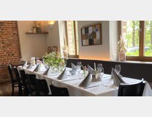 Langer, stilvoll gedeckter Tisch in einem gemütlichen Raum mit Backsteinwand, weißen Tischdecken und Gläsern. Große Fenster lassen Tageslicht herein, dekoriert mit Blumenarrangements und dezentem Wandschmuck.