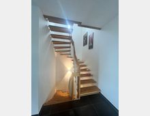 Eine moderne Innentreppe aus Holz mit offenen Stufen führt in einem weißen Treppenhaus mit schwarzen Bodenfliesen nach oben und unten. An den Wänden hängen zwei gerahmte Bilder, und eine Wandlampe strahlt warmes Licht aus.