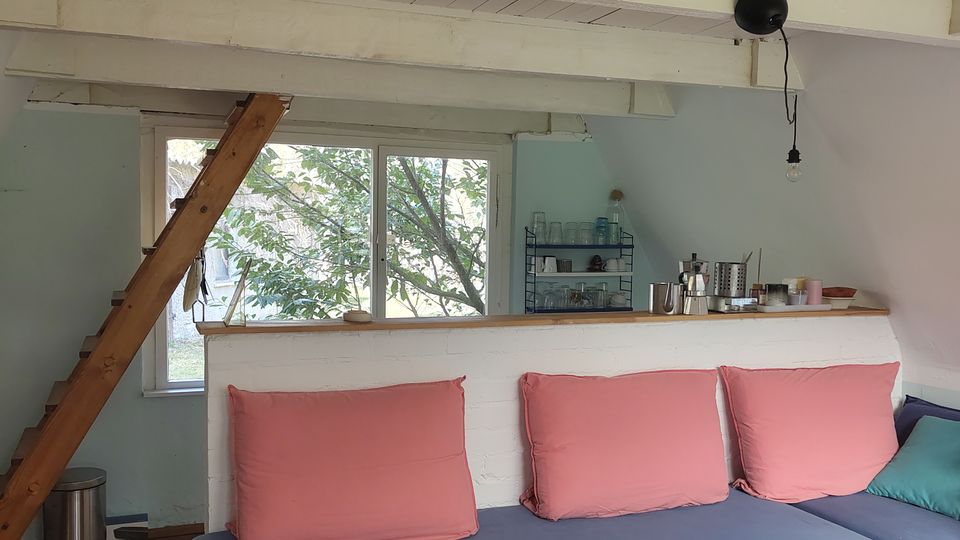 Innenraum eines kleinen Dachgeschoss-Studios mit weiß gestrichenen Holzbalken und niedrigem Plafond, großem Fenster und einer einfachen Küchenzeile im Hintergrund. Im Vordergrund steht ein Paletten-Sofa mit blauem Polster und rosa Kissen auf Holzboden, daneben eine schräge Holztreppe und eine hängende Lampe.