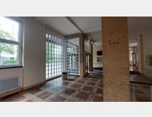 Innenraum eines Gebäudes mit großen Glasbausteinfenstern, einer tragenden Ziegelsäule und einem Boden aus bunten Fliesen. Im Hintergrund sind Türen, eine grüne Notausgangsleuchte und ein Teil eines offenen Flurs zu sehen.