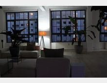 Großzügiger Loft-Wohnraum mit großen, dunklen Sprossenfenstern, die auf ein modernes Wohngebäude blicken. Im Vordergrund befinden sich helle Sofas, Zimmerpflanzen in Töpfen und eine Stehlampe mit warmem Licht.