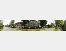 Panorama eines historischen Dorfplatzes mit Kopfsteinpflasterstraßen, umgeben von alten Gebäuden mit roten Ziegeldächern und Putzfassaden. Die Szene ist von hohen Bäumen und einer grünen Umgebung geprägt.