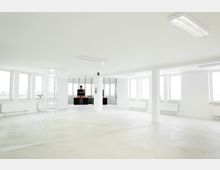 Ein nahezu leerer, heller Innenraum mit weißen Wänden, Deckenleuchten, großen Fenstern und einem hellen Boden. Es gibt mehrere Heizkörper unter den Fenstern und im Hintergrund ist ein kleiner Bereich mit schwarzen Regalen und Elektronik zu sehen.