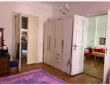 Ein Schlafzimmer in einer Altbauwohnung mit Holzdielenboden und Stuck an der Decke. Im Raum stehen ein großer weißer Kleiderschrank, ein Holztisch, und rosa Bettwäsche liegt auf dem Bett; durch zwei geöffnete Türflügel sind ein angrenzendes Wohnzimmer mit roten Sitzmöbeln und eine Küche sichtbar.