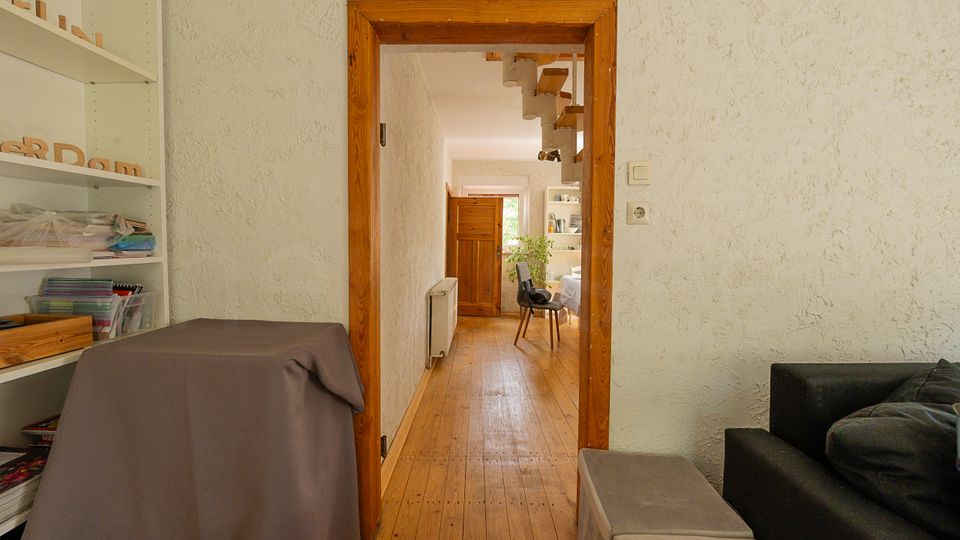 Blick durch einen holzgerahmten Türdurchgang in einen schmalen Flur mit Holzfußboden und weiß strukturierten Wänden. Links stehen Regale, am Ende führt der Flur in einen hellen Raum mit Tisch, Stuhl und Fenster.