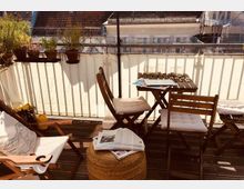 Eine kleine Dachterrasse mit Holzboden, ausgestattet mit mehreren Holzklappstühlen und einem kleinen Tisch. Im Hintergrund sind Blumentöpfe auf einem Geländer sowie Dächer umliegender Häuser sichtbar.