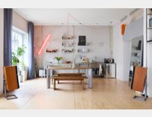 Minimalistisch gestaltete Wohnküche mit Holzboden, Betonwand und offenem Regalsystem. Ein Holztisch mit Bänken steht im Vordergrund, während im Hintergrund eine Edelstahlküchenzeile mit Spüle, Kochutensilien und roten Akzenten zu sehen ist, flankiert von großen Fenstern mit bodenlangen Vorhängen.