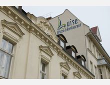 Fassade eines historischen Gebäudes mit dekorativen Stuckelementen und Fenstern mit Dreiecksgiebeln; auf der oberen rechten Wand ist das Logo „Luise Bistro & Restaurant“ angebracht.