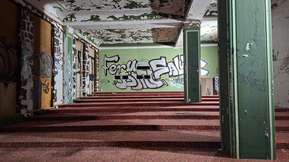 Innenraum eines verlassenen Gebäudes mit breiten, rotbraunen Stufen, grünen Wänden und mehreren Stützpfeilern. Abblätternde Deckenfarbe, teils zugemauerte Fenster und großflächige Graffiti prägen den Raum.