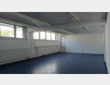 Ein leerer, rechteckiger Raum mit einem dunkelblauen Boden, weißen Wänden und einer abgehängten Decke mit integrierten rechteckigen Leuchtstofflampen. Große Fenster an einer Wand lassen Tageslicht herein, während darunter Heizkörper und Rohre sichtbar sind.