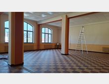 Großer, leerer Innenraum mit kariertem Fliesenboden, mehreren tragenden Säulen und einer abgehängten Rasterdecke. An der Außenwand befinden sich große Rundbogenfenster mit Heizkörpern darunter; rechts steht eine aufgeklappte Stehleiter.