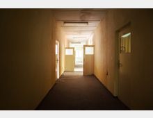 Ein leerer, schlecht beleuchteter Flur mit Teppichboden, beige Wänden und mehreren Türen mit Glasfenstern, teilweise offen. Am Ende des Flurs ist ein Fenster erkennbar, das Tageslicht hereinlässt.