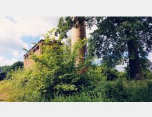 Industriegelände mit einem verlassenen Backsteingebäude und einem hohen, mit Graffiti versehenen Schornstein, umgeben von dichter Vegetation und Bäumen. Der Himmel ist teilweise bewölkt, und das Gelände wirkt überwuchert.