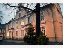 Historisches Gebäude mit heller Fassade und hohen Fenstern, darunter zwei Eingangsportale mit kleinen Vordächern; im Vordergrund steht ein kahler Baum und ein beleuchteter Tannenbaum. Die Umgebung zeigt einen gepflasterten Platz mit Fahrrädern und Bänken bei einsetzender Dämmerung.