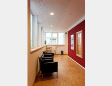 Ein modern eingerichteter Flur mit hellem Holzfußboden, großen Fenstern auf der linken Seite und einer roten Wand auf der rechten Seite. Der Raum ist mit schwarzen Sesseln, einem kleinen Tisch mit Hockern sowie dekorativen Bildern ausgestattet.