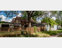 Ein ländliches Grundstück mit einem alten Backsteinhaus, einer angrenzenden Garage und mehreren Garagenboxen. Im Vordergrund steht ein Holzlattenzaun, daneben große Bäume und grüne Vegetation, die einen schattigen Weg säumen.