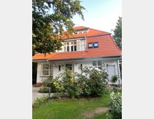 Zweistöckiges Wohnhaus im traditionellen Stil mit weißer Fassade und rotem Ziegeldach, umgeben von gepflegtem Garten mit Sträuchern und Bäumen. Ein gepflasterter Weg führt zum Eingangsbereich, der von einem kleinen Vordach geschützt ist.