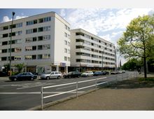 Mehrstöckige Wohn- und Geschäftshäuser mit weißen Fassaden an einer belebten Straße, vor denen Autos parken. Im Erdgeschoss der Gebäude befinden sich Geschäfte, darüber befinden sich Balkone und Fenster; ein Baum steht rechts im Bild.