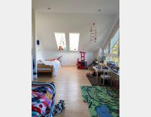 Ein kindgerecht eingerichtetes Zimmer mit Dachschrägen, zwei Dachfenstern und hellem Holzfußboden. Es enthält ein Bett, Spielzeug, eine Spielküche, einen Tisch mit Stühlen und einen bunten Spielteppich mit einem Stadtmotiv.