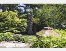 Japanischer Garten mit üppigem Grün, darunter Bambus und Bäume, sowie einem großen Stein mit goldenen Schriftzeichen. Im Vordergrund sind Steine und Gras sichtbar, die den Weg durch den Garten säumen.