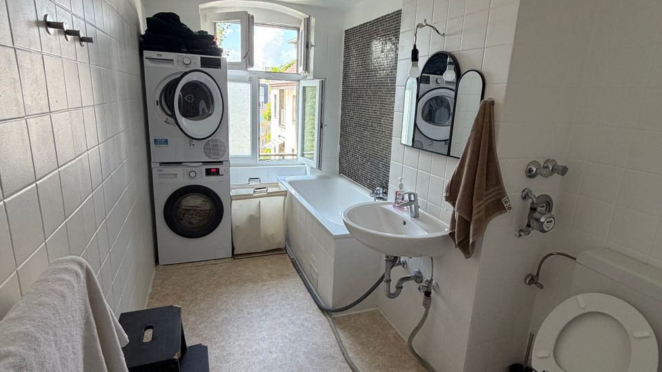 Schmales Badezimmer mit weißen Fliesen, Waschbecken mit Spiegel, Toilette und Badewanne unter einem großen Fenster. Am Fenster steht eine gestapelte Waschmaschine mit Trockner; links ist ein Heizkörper und ein kleiner Hocker, rechts ein dunkler Fliesenstreifen als Akzentwand.