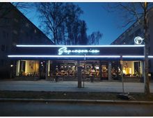 Straßenansicht eines Restaurants mit moderner Fassade und leuchtendem Schriftzug „Supersonico“ in Neonlicht. Im Innenraum sind Beleuchtung, Möbel und Dekoration erkennbar, umgeben von Bäumen und Wohnhäusern im Hintergrund bei Dämmerung.