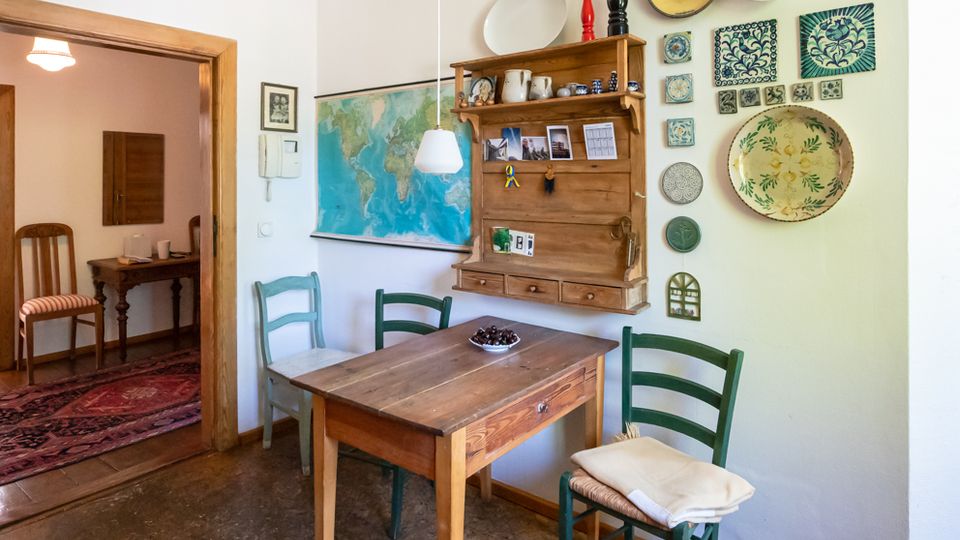 Gemütlicher Essbereich mit kleinem Holztisch und drei Stühlen, einer hängenden Lampe und einer großen Weltkarte an der Wand. Daneben hängen ein rustikales Wandregal mit Keramik sowie dekorative Teller und Fliesen; durch einen Holztürrahmen ist ein angrenzendes Zimmer mit Teppich zu sehen.