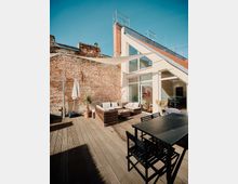 Dachterrasse mit Holzboden, ausgestattet mit einer Sitzgruppe aus Rattanmöbeln und einem Essbereich mit schwarzem Tisch und Stühlen. Im Hintergrund eine Fassade aus Ziegelsteinen und eine moderne Glasfront mit schrägem Dach, darüber ein schattenspendendes Sonnensegel.