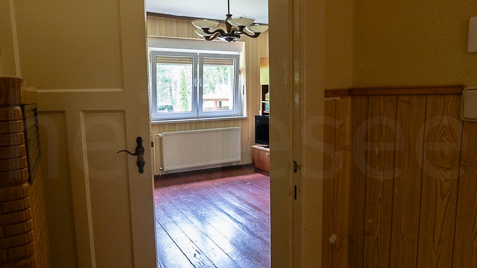 Blick durch eine geöffnete Tür in einen älteren Wohnraum mit dunklem Holzdielenboden, Fensterfront und Heizkörper darunter. Im Vordergrund ist ein schmaler Flur mit Holzvertäfelung zu sehen, im Raum hängt ein Deckenleuchter.