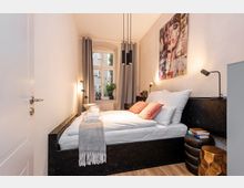 Schmales, modernes Schlafzimmer mit Doppelbett in dunklem Bettrahmen, weißer Bettwäsche und grauem Plaid sowie Nachttischen mit Leselampen. Am Ende des Raums steht ein großes Fenster mit grauen Vorhängen; darüber hängen schwarze Pendelleuchten und an der Wand ein großes Porträtbild.