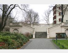 Treppenaufgang mit Balustraden, flankiert von symmetrischen Mauern mit Rundbögen, in einem Park mit gepflastertem Weg, Grünflächen und kahlen Bäumen; im Hintergrund ein mehrstöckiges Gebäude.