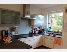 Moderne Küche mit weißen Schränken und dunkler Arbeitsplatte, ausgestattet mit Dunstabzugshaube, Wasserkocher, Kaffeemaschine und weiteren Küchengeräten. Ein großes Fenster bietet Blick nach draußen, und auf der Ablage stehen frische Lebensmittel und Dekorationen wie eine Orchidee.