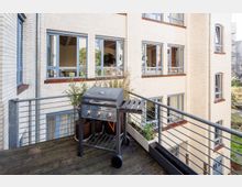 Ein Balkon mit Holzboden und Metallgeländer, auf dem ein Gasgrill steht. Im Hintergrund ist die helle Fassade eines mehrstöckigen Gebäudes mit großen Fenstern sichtbar, umgeben von etwas Grün.