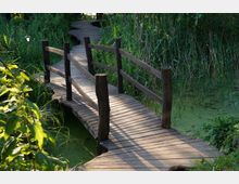 Holzsteg mit Geländer, der über grüne, mit Pflanzen bewachsene Gewässer führt, umgeben von hohen Gräsern und dichten Bäumen in einer natürlichen, bewaldeten Umgebung. Eine Laterne steht links neben dem Steg.