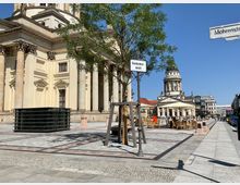 Blick auf den Gendarmenmarkt in Berlin, mit dem Französischen Dom im Vordergrund und dem Deutschen Dom im Hintergrund. Der Platz zeigt Sitzbereiche mit Sonnenschirmen, Bäume sowie gepflasterte Gehwege, umgeben von klassizistischer Architektur.