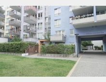 Mehrstöckiges Wohngebäude mit hellblauer und beiger Fassade, mehreren Balkonen und einem Durchgang zu einem Innenhof. Im Vordergrund sind eine kleine Rasenfläche, Hecken und ein Zaun zu sehen.