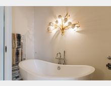 Helles Badezimmer mit freistehender, weißer Badewanne und einer dekorativen goldfarbenen Wandleuchte in floralem Design. Im Hintergrund ein Handtuchhalter mit gemusterten Handtüchern an einer cremefarbenen Wand.